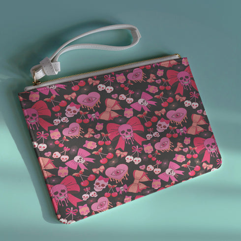Rectangular wristlet clutch featuring a dark background with pink skull bows, cherries, and other goth-inspired motifs. White strap and gold zipper visible.